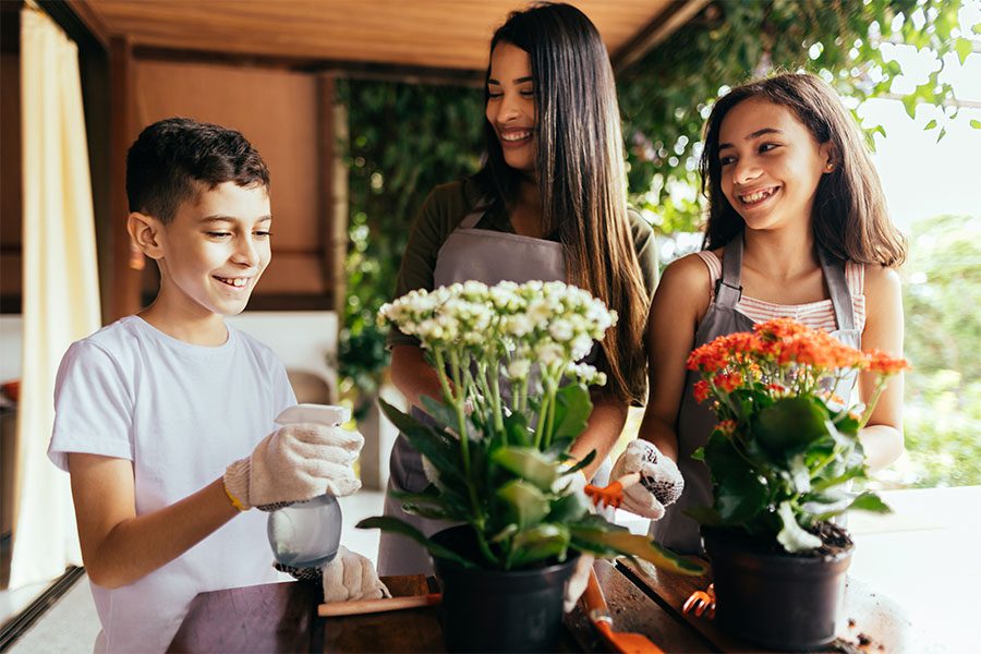 About Our Agency - Portrait of a Young Mother and Her Two Kids Having Fun Spending Time Together Outside on the Deck in the Backyard While Taking Care of Flowers
